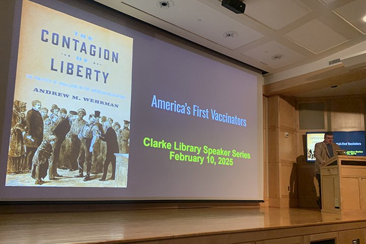 Andrew Wehrman stands behind a wooden podium on a stage with his book cover projected on a large screen next to him.