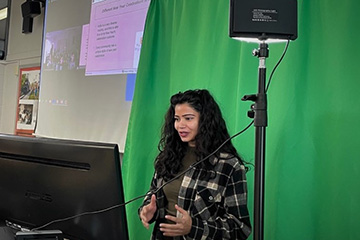 A CMU student standing in front of a green screen presents a live virtual field trip session for the Museum of Cultural and Natural History.