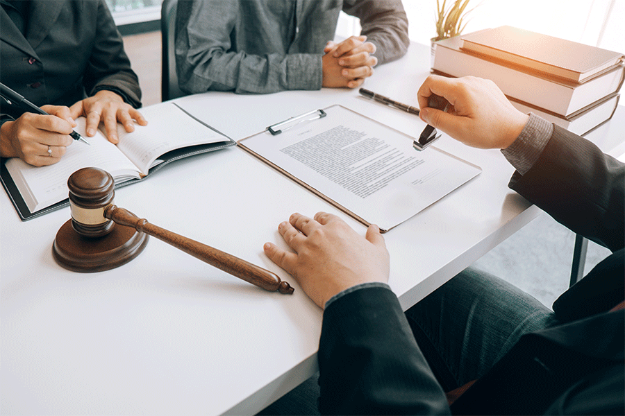 Three individuals are seen sitting at a white table with a gavel next to one, and documents and books are on the table.