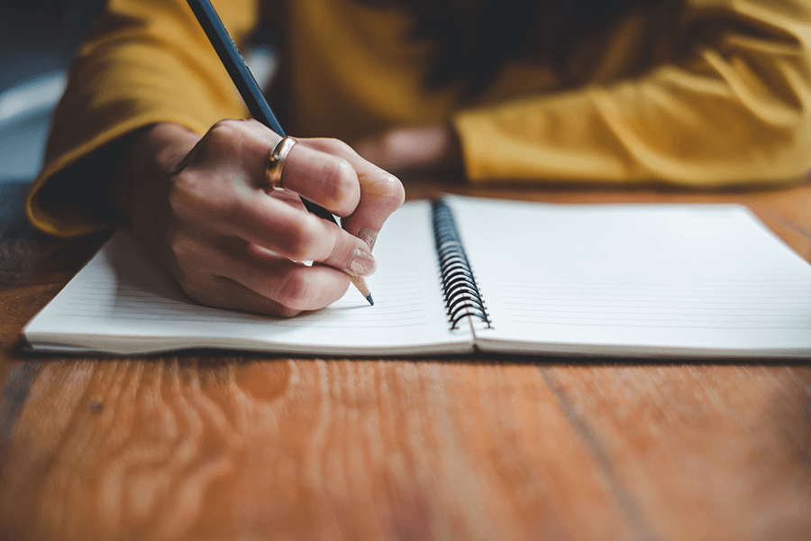 A person wearing a mustard-yellow shirt holds a pencil to a blank notebook on a wooden desk as if about to write.