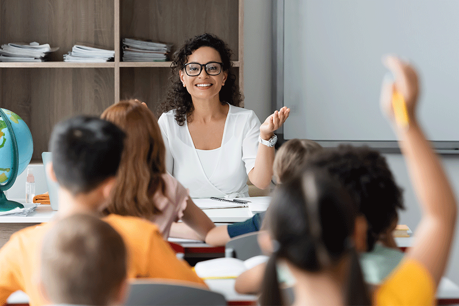 An elementary school teacher sitting in a classroom, facing young students with their hands raised as she calls on one.