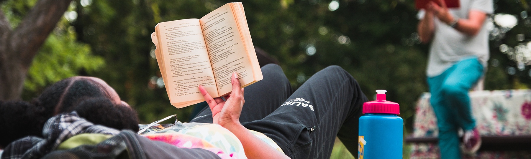 English literature student wearing black sweatpants and a tie-dye shirt lays back on the grass while holding a book up and reading during an outdoor class.