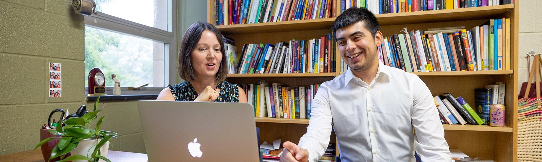 English faculty member Carlin Borsheim-Black and student Marcelino Olivarez view laptop screen while seated in front of bookshelves filled with books.