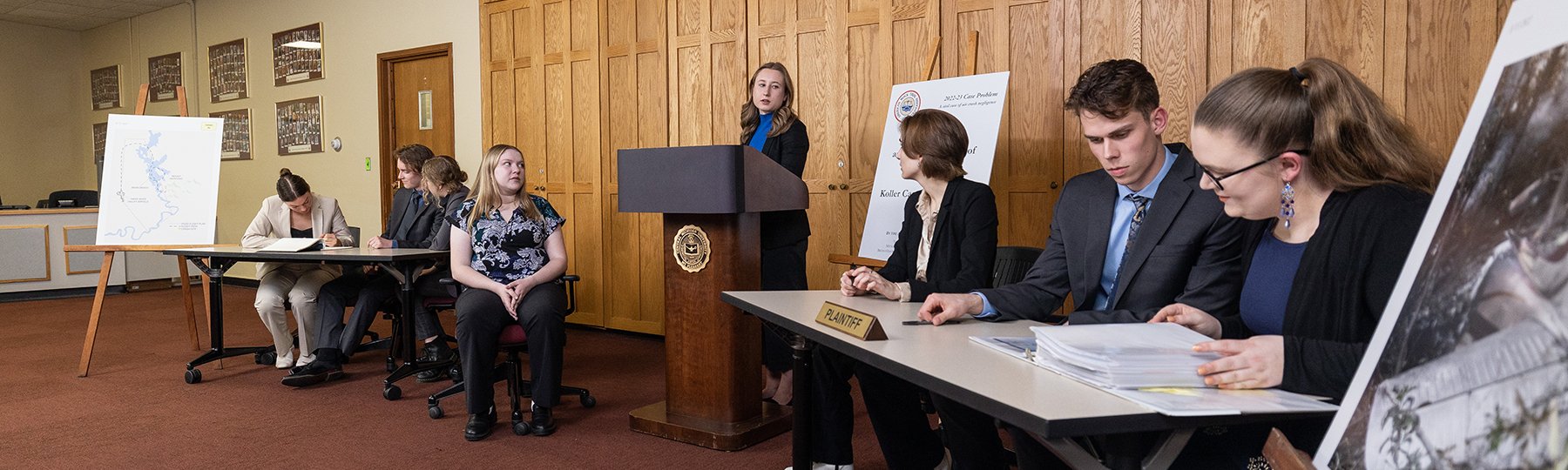 Students question a witness seated next to tables and a podium during a mock trial presentation.