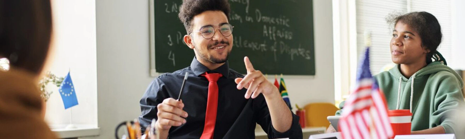 A teacher wearing a black shirt and red tie sits in a social studies classroom with students.