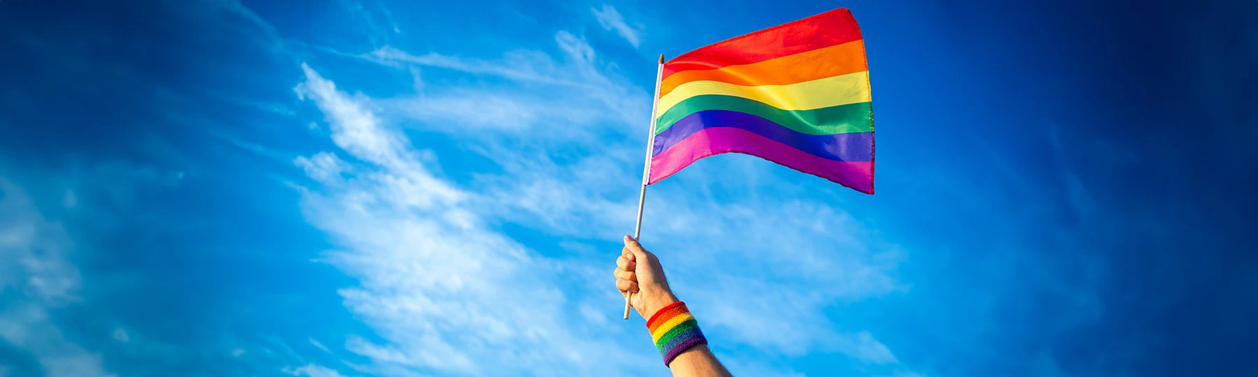 Image of a Pride Flag blowing in the wind on a flagpole with a blue sky in the background.