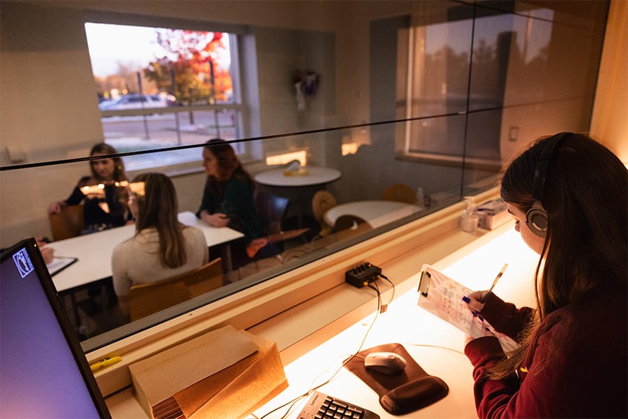 An experimental psychology student takes notes from behind a one-way mirror as a group meets and talks.