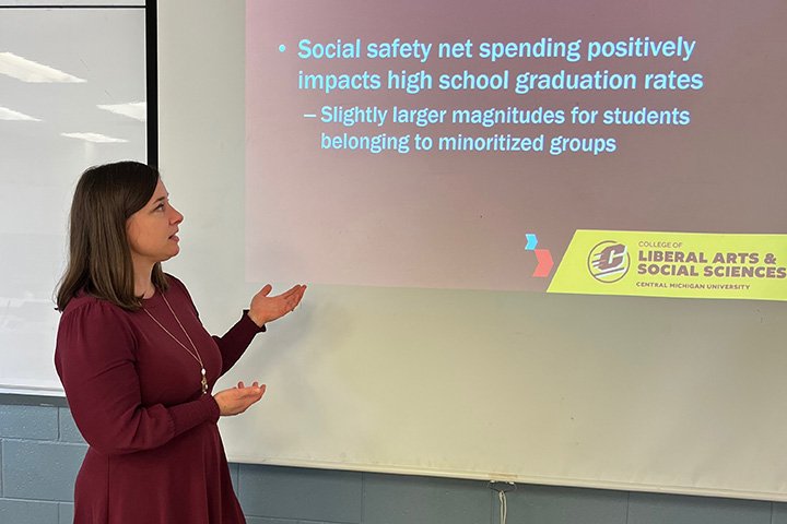 Faculty member Sharon Kukla-Acevedo stands next to a digital display board featuring text about social safety net spending and high school graduation rates.