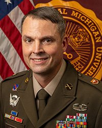 Closeup image of Master Sergeant Charles “Chuck” Lee wearing military uniform and seated in front of an American flag and the Central Michigan University seal.