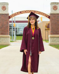 Ava Brewer in a Maroon Cap and Gown standing underneath a brick archway that says Central Michigan University.
