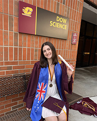 Claire DeBlanc wearing a while dress with a maroon gown holding a rolled up diploma in front of a brick wall with a sign that says Dow Science.