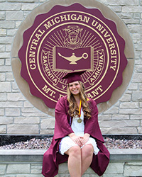 Meghan VanDamme in a white dress and maroon cap and gown sitting on a brick ledge in front of the Central Michigan University seal.