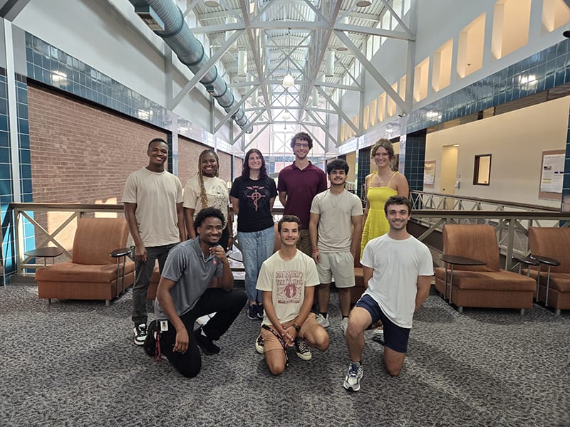 A group of students posing together with the interior of the Engineering and Technology building behind them.