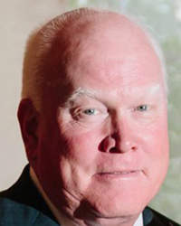 A man with white hair wearing a suit and tie looks into the camera.