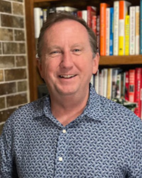 A man wearing a blue patterned button up stands in front of a book shelf.