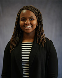 A woman in a striped shirt and black jacket in front of a grey backdrop.