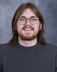 A young man with shoulder-length brown hair, a beard, and glasses, wearing a gray sweater. He is smiling against a neutral gray backdrop.