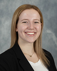 A woman with long brown hair wearing a white shirt and a dark blazer smiling at the camera in front of a grey backdrop.