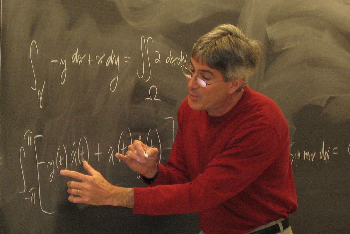 A man in a red shirt and glasses standing in front of a chalk board pointing at a complex mathematical equation.