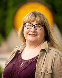 A woman in glasses and a brown jacket smiling at the camera.