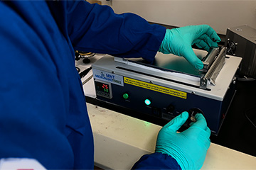 A person in a blue lab coat and gloves applies a black material to a metal plate.