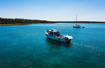 The MV CMU Chippewa moves through clear turquoise waters, leaving a gentle wake behind it. In the background, a sailboat is anchored near a small dinghy, with a tree-lined shoreline stretching across the horizon under a bright blue sky.