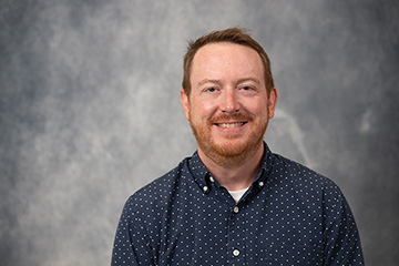 Marty Baxter in a blue button up shirt standing in front of a grey backdrop.