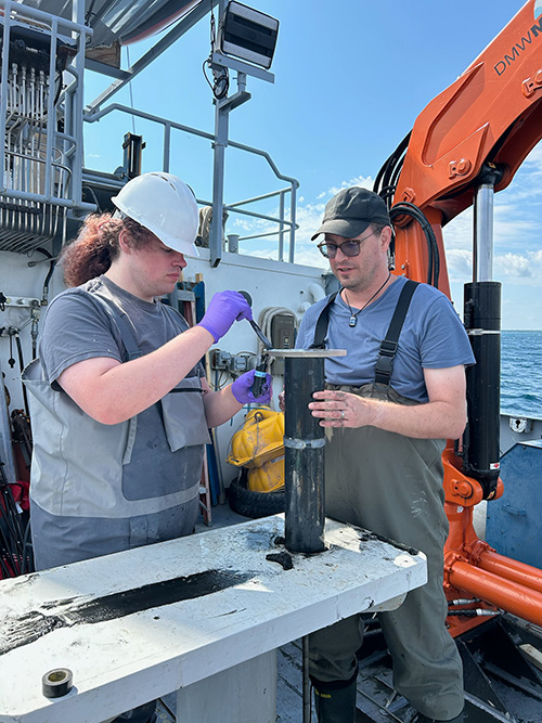 Carmine Sabatini in a grey shirt, hard hat, and grey waders taking samples on the deck of a ship with Anthony Chappaz in a blue shirt, hat, and waders.