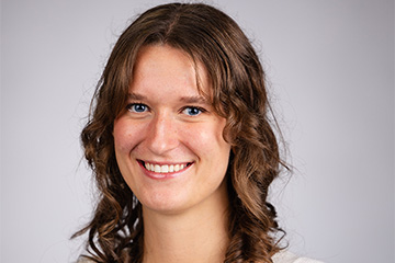 A woman with blue eyes and curly hair smiling at the camera in front of a grey backdrop.