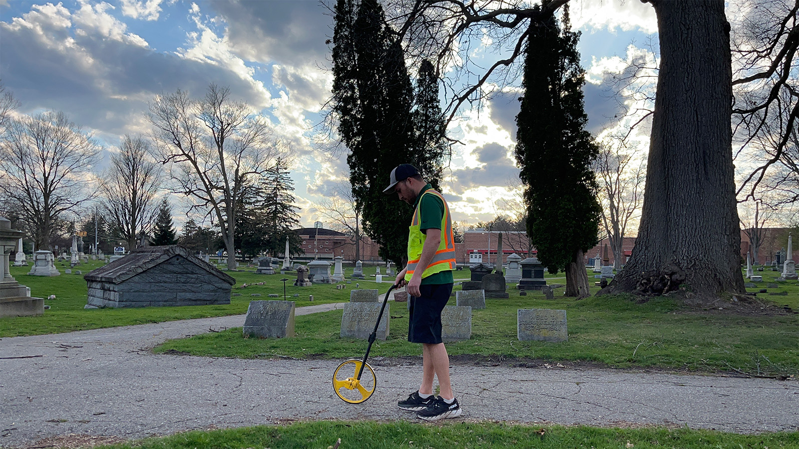 Anthony Kowalchick in a cemetery, wearing shorts and a high visibility vest using a rolling device to measure ground distance.