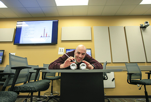 Man in a classroom setting leans over a table with two aluminum cans, appearing engaged in a demonstration or experiment. The background features a presentation screen displaying a graph and text, along with acoustic panels on the walls.