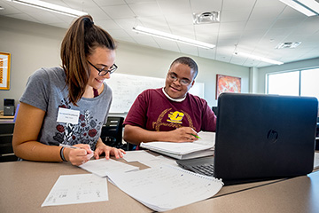 Two students at a table reviewing mathematics homework with a laptop and papers in front of them.