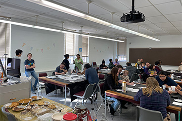 A classroom filled with people of various ages engaging in activities at long tables covered with papers.