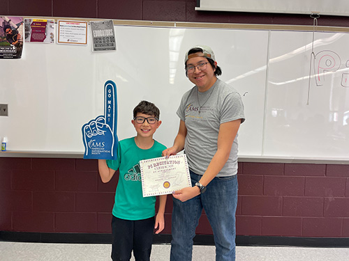 A young student wearing glasses and a green t-shirt holds up a blue foam finger while standing next to an adult wearing a gray t-shirt and a backward cap. The student is smiling and holding a certificate.