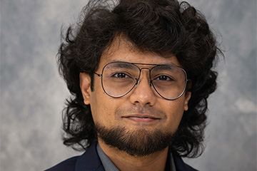 A man with dark hair, glasses and beard in front of a grey backdrop.