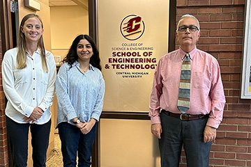 A woman with long blonde hair, a woman with long dark hair, and a man with glasses and a red shirt standing in front of a sign that says CMU School of Engineering and Technology.