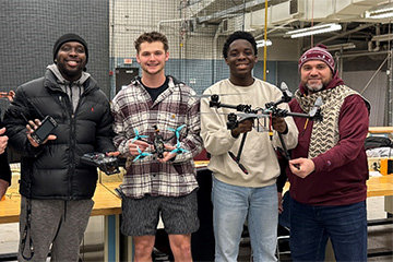 Four members of CMU’s D.A.R.E (Drone Aviation, Research, and Engineering) student organization pose together in a workshop, proudly displaying their drones.