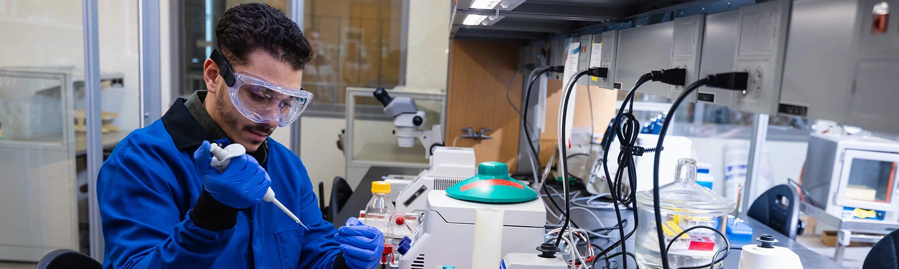 A student wearing safety glasses, a lab coat and gloves works with a pipette in a biotechnology lab on campus.