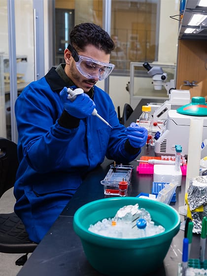 A student wearing safety glasses, a lab coat and gloves works with a pipette in a biotechnology lab on campus.