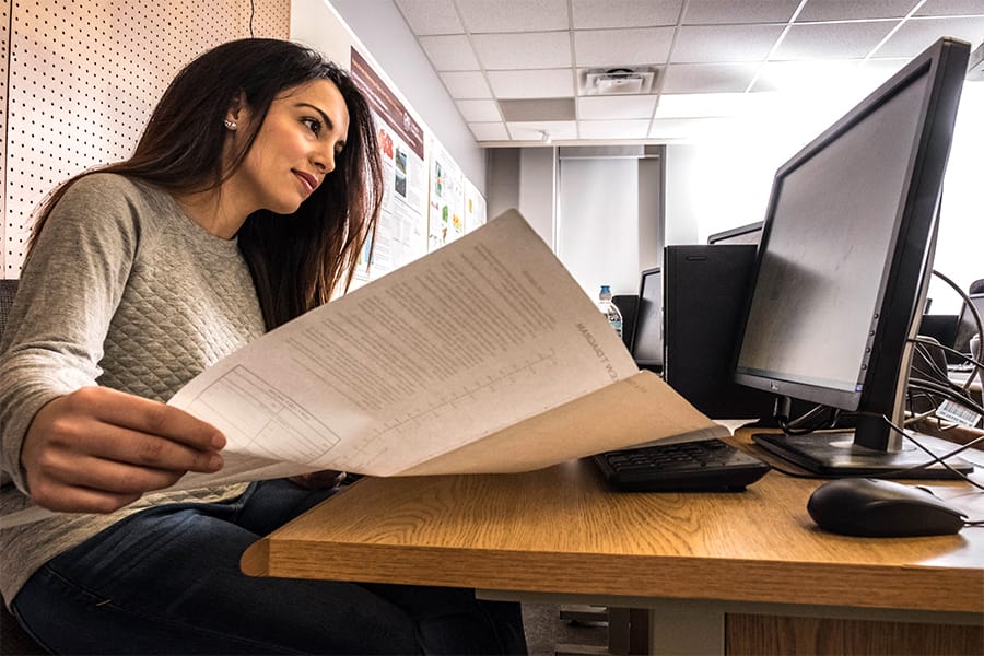 A woman with long dark hair and a light-colored blouse holds a piece of paper open in front of a computer screen.