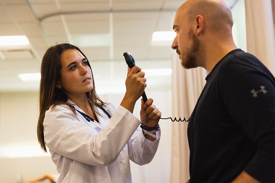 A pre-optometry student holds a tool used to look at eyes up to a male patient sitting on a bed.