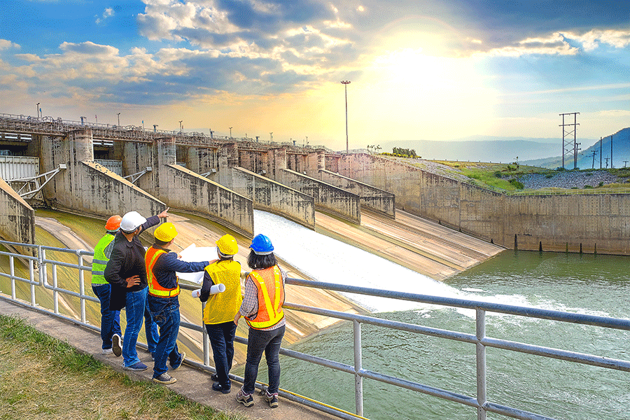 Four environmental engineers stand against a bridge and look out to a water distribution center as they wear hard hats and neon construction vests.