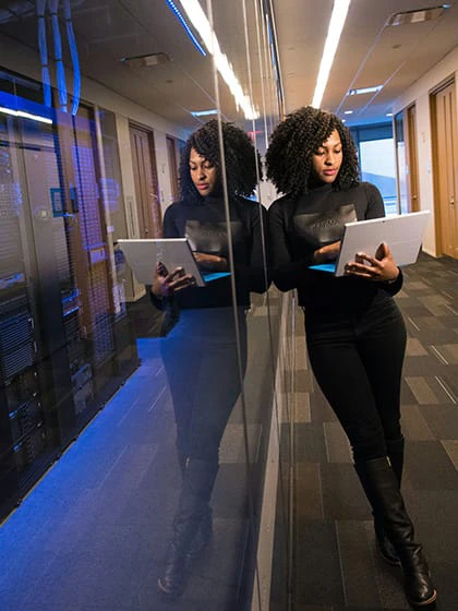 A woman with dark curly hair dressed in all black and holding a laptop leans against a glass panel on the outside of a server room.