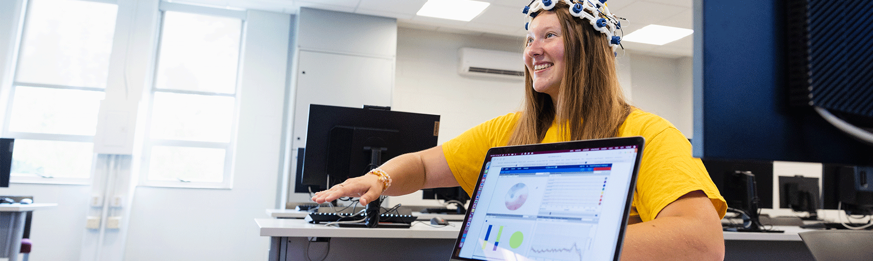 A student sitting in a classroom, with a tool on her head that is a white cap with blue dots, as she raises her hand and a computer is on the table in front of her showing charts and data.