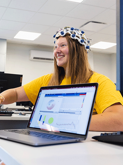 A student sits in a classroom with a research tool on her head and a computer is displaying data for a computer science degree project.