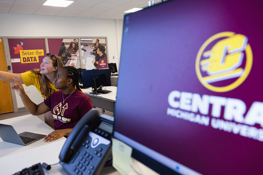 Two data science students wearing maroon and gold, pointing and looking at a screen in a classroom, with a laptop in front of them. The wall behind them has a large sticker that reads. "Seize the data". There is another computer screen with an action C.