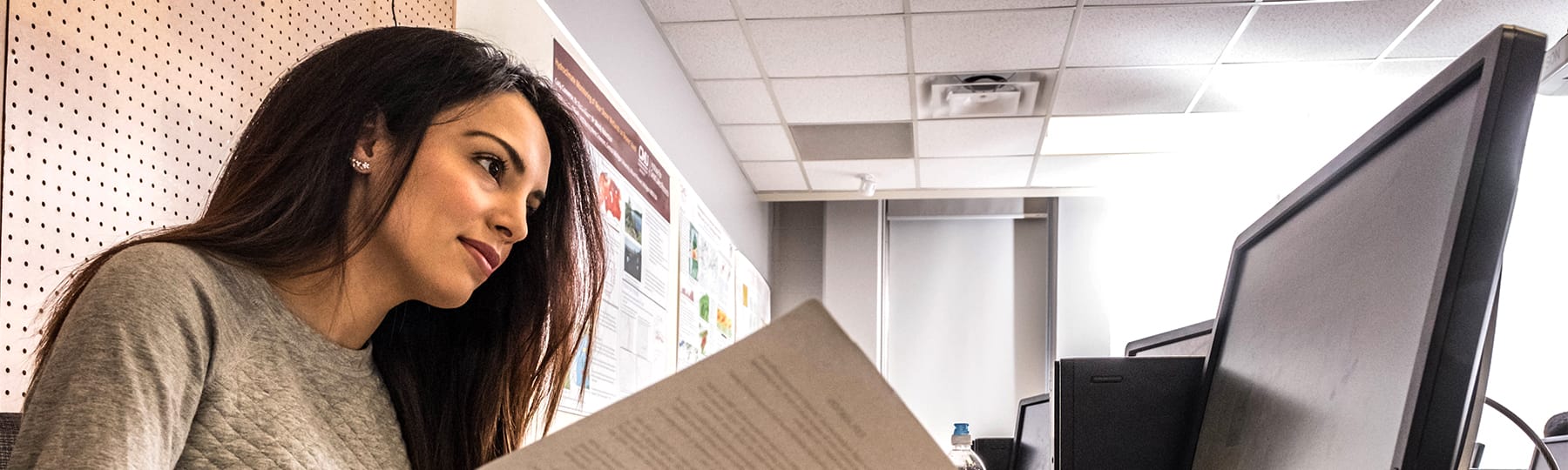 A woman with long dark hair and a light-colored blouse holds a piece of paper open in front of a computer screen.