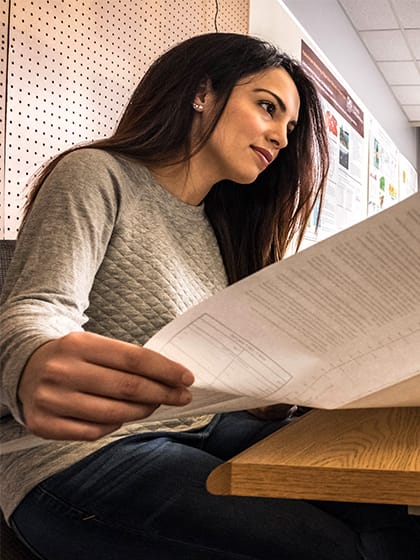 A woman with long dark hair and a light-colored blouse holds a piece of paper open in front of a computer screen.