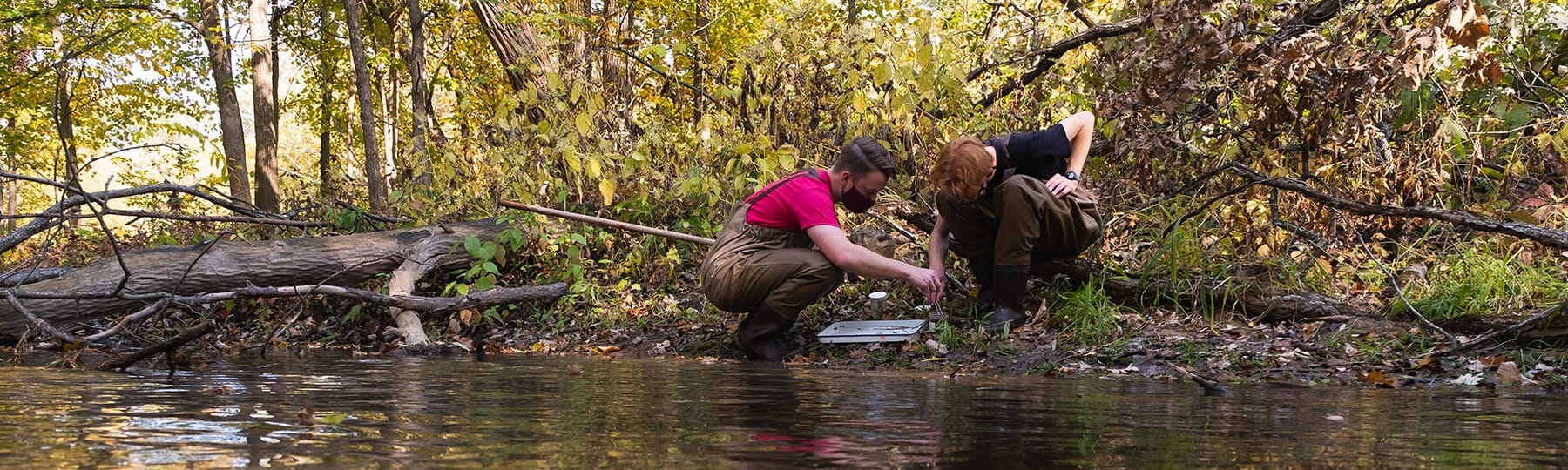 Two environmental studies students crouch on the bank of a river to examine a sample.