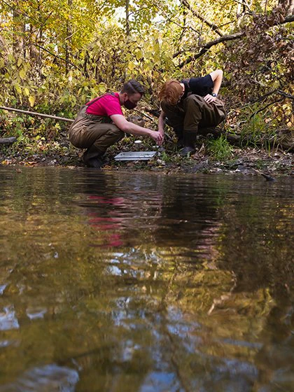 Two environmental studies students crouch on the bank of a river to examine a sample.